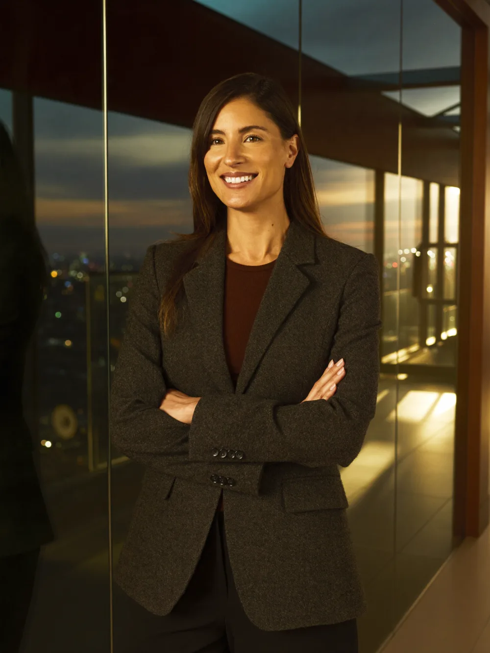 Smiling team lead with arms crossed beside a glass wall at dusk